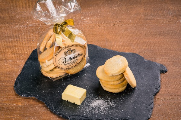 Butterspritzgebäck in durchsichtiger Geschenkverpackung mit goldener Schleife neben einem Stück Butter und Puderzucker auf Schieferplatte.